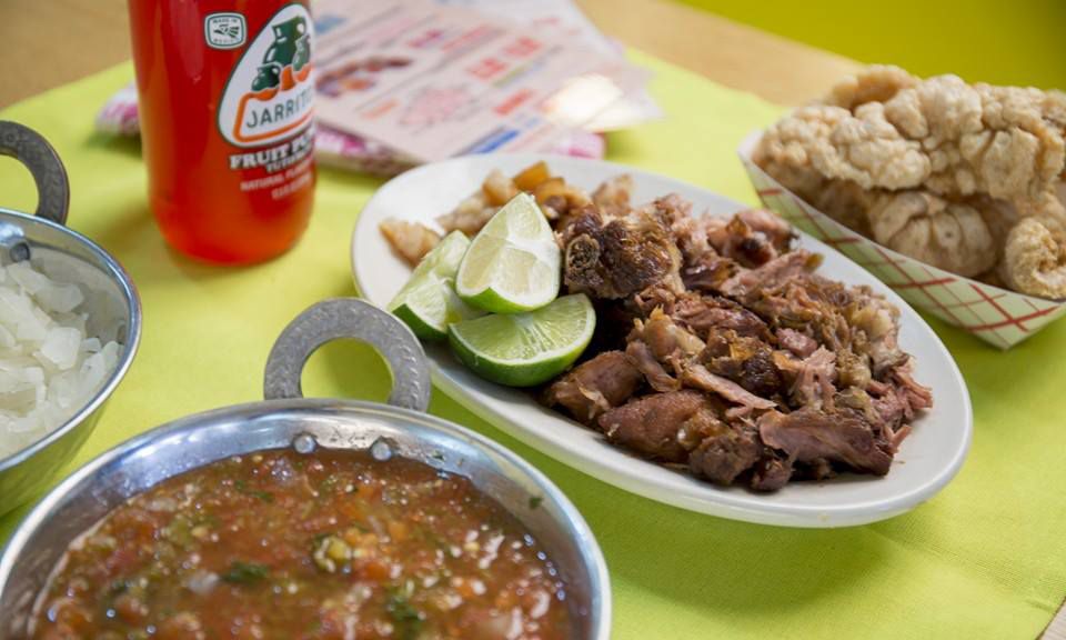 A platter of meat, a bowl of salsa, a paper container of chicharron, and a red Jarritos soda sit on a green tablecloth.