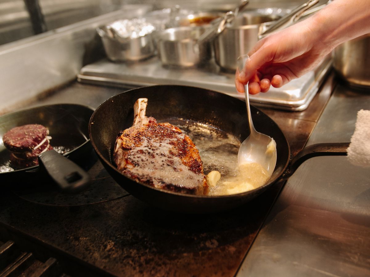 A chef bastes a steak in butter on a stove.