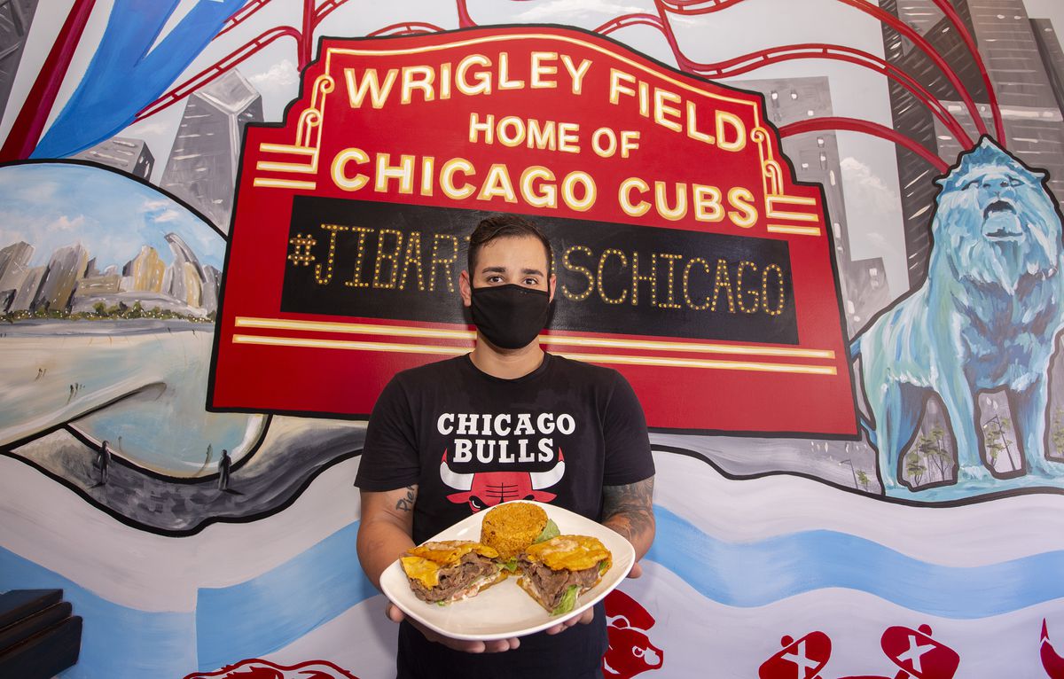 A man in a black mask and black Chicago Bulls t-shirt stands in front of a wall painted with a mural of the big red Wrigley Field sign holding a plate of jibaritos.