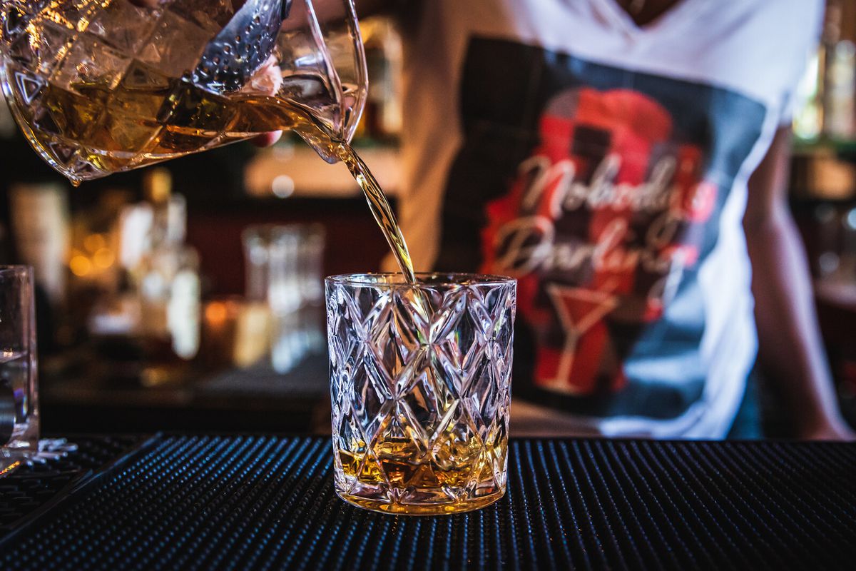 A bartender pours brown liquor into a fancy rocks glass
