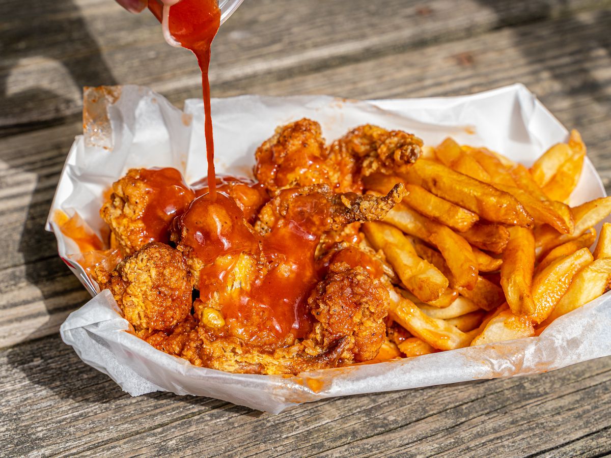 Red sauce drizzles from a cup over a basket of fried chicken and fries.