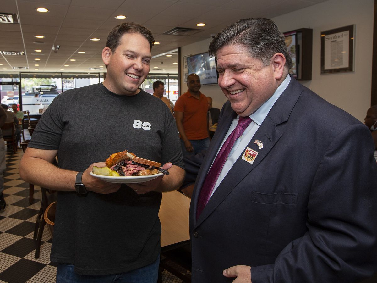 Two smiling men, one holding a giant pastrami sandwich.