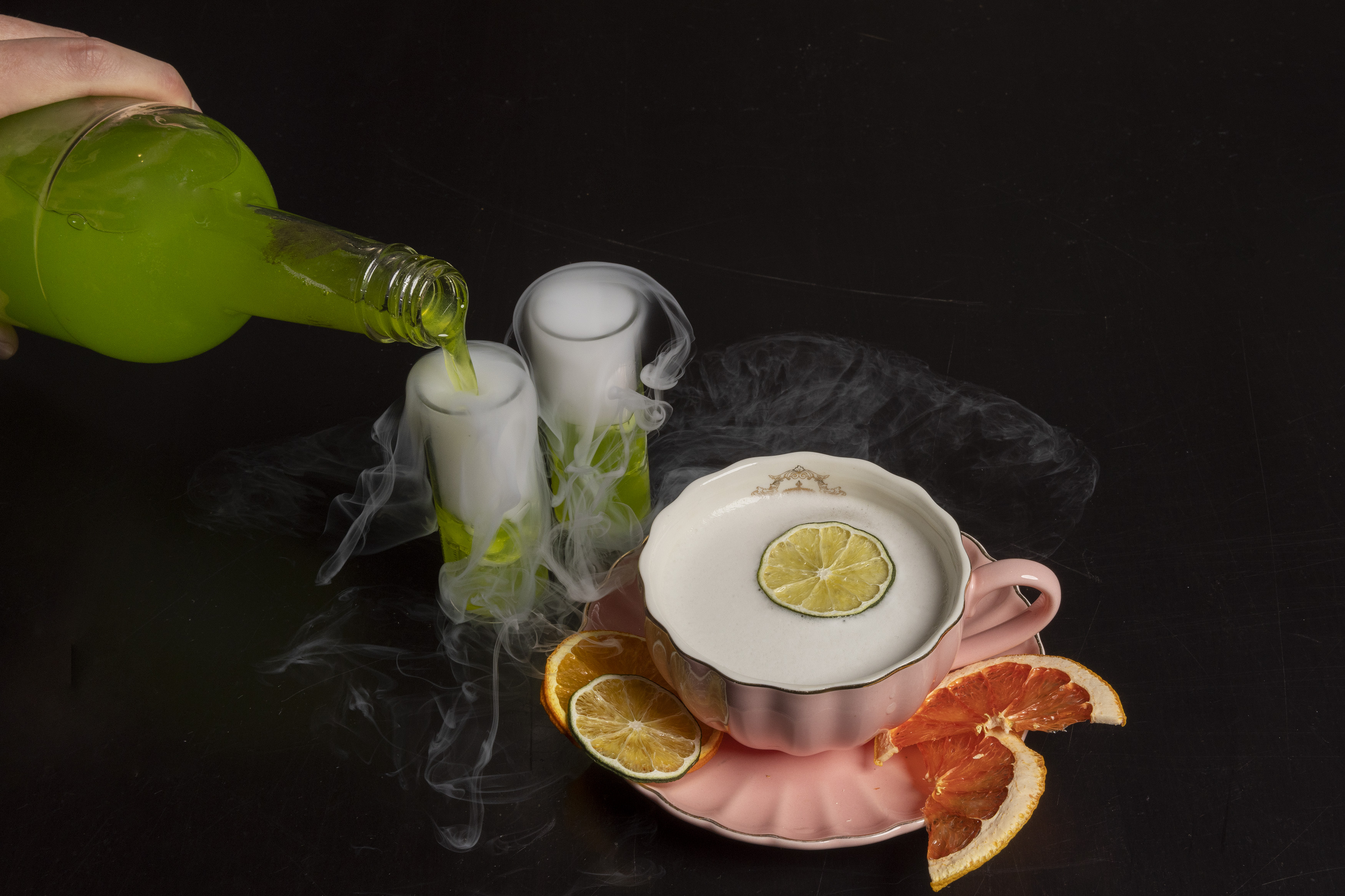 A hand pours green liquid into a smoking shot glass beside a pink teacup.