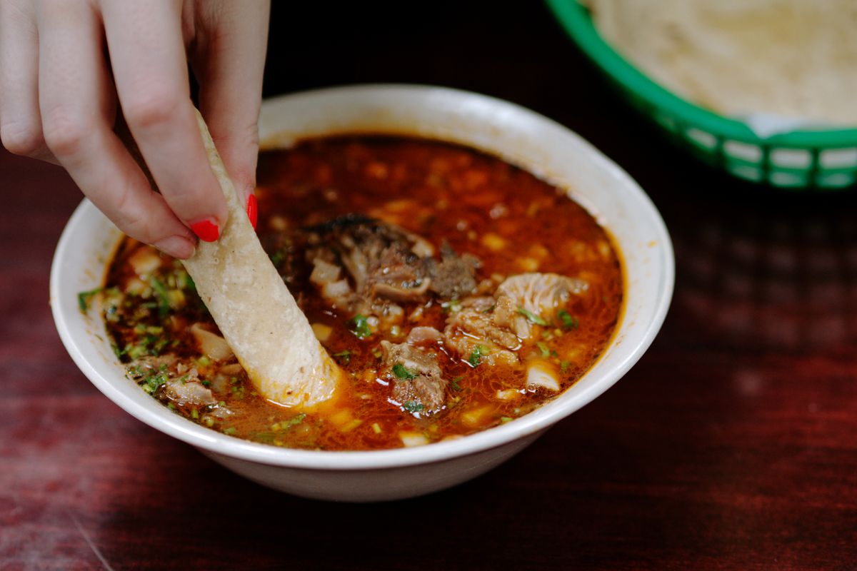 A hand dips a rolled tortilla into a bowl of birria.