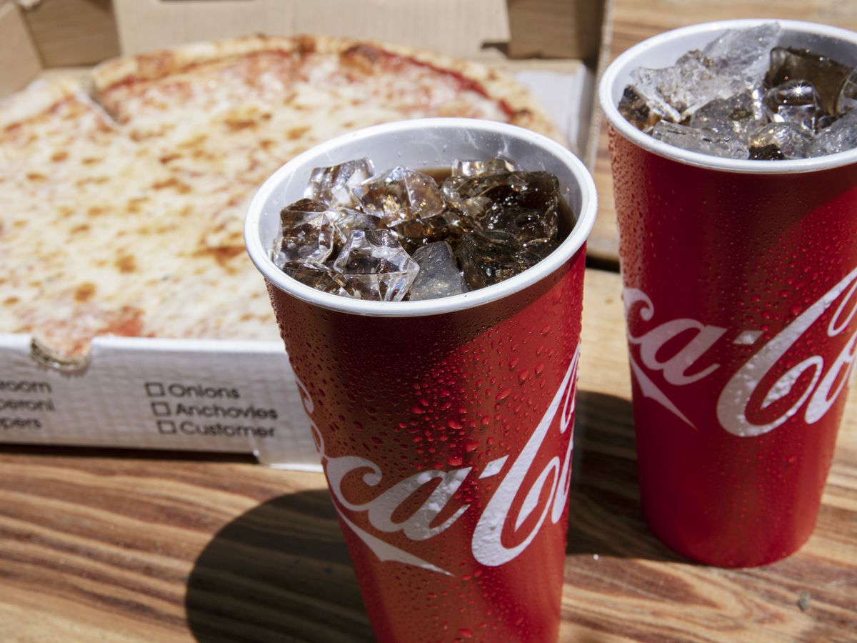 A cheese pizza in a delivery box, plus two paper cups of Coca-Cola.