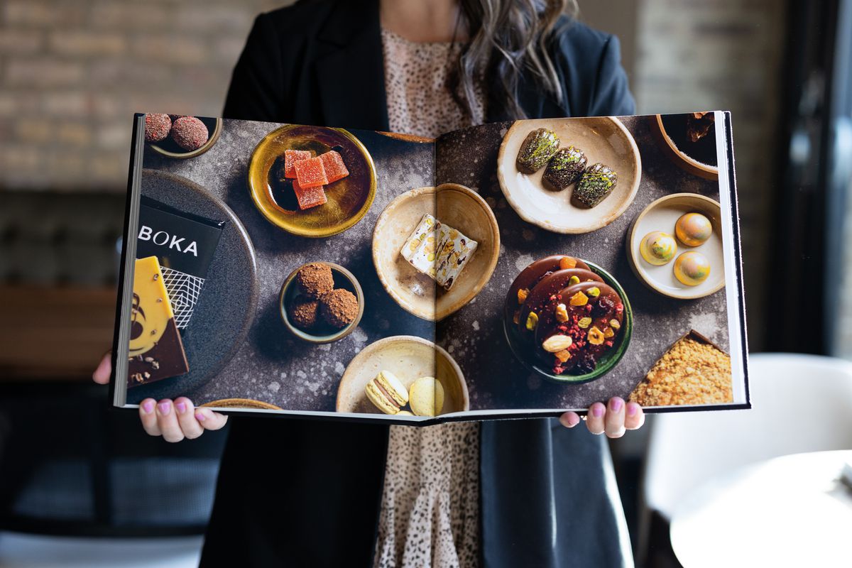 A person holds a large cookbook open to a full-bleed photograph of food on a table.