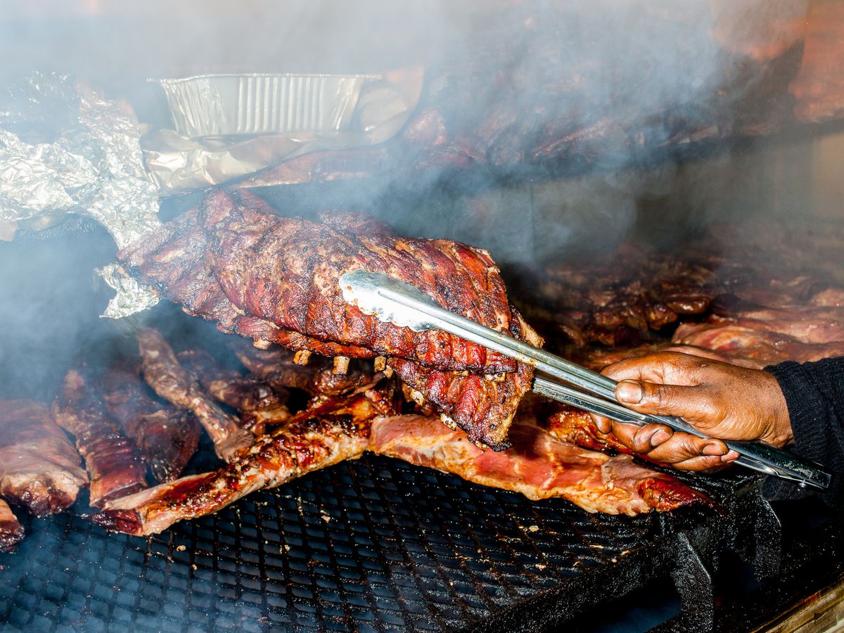 A hand holds aloft a rack of ribs with a tong in front of a full smoker.