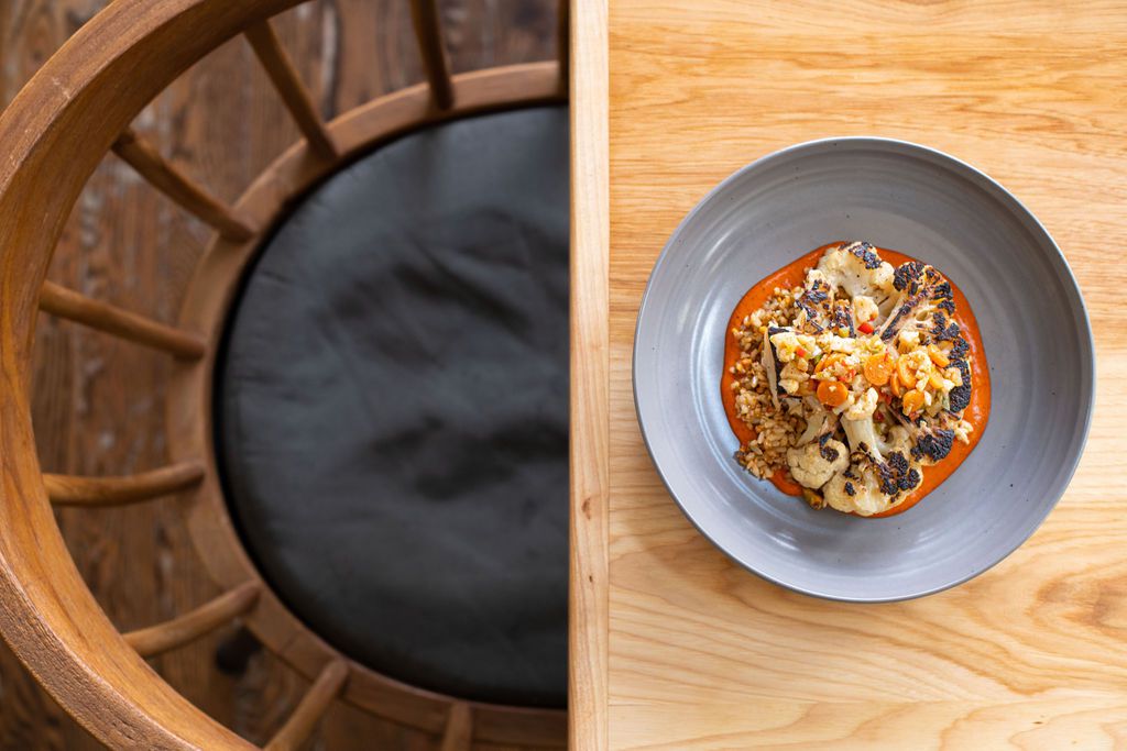 A colorful cauliflower dish sits inside a grey bowl on a light wood table.
